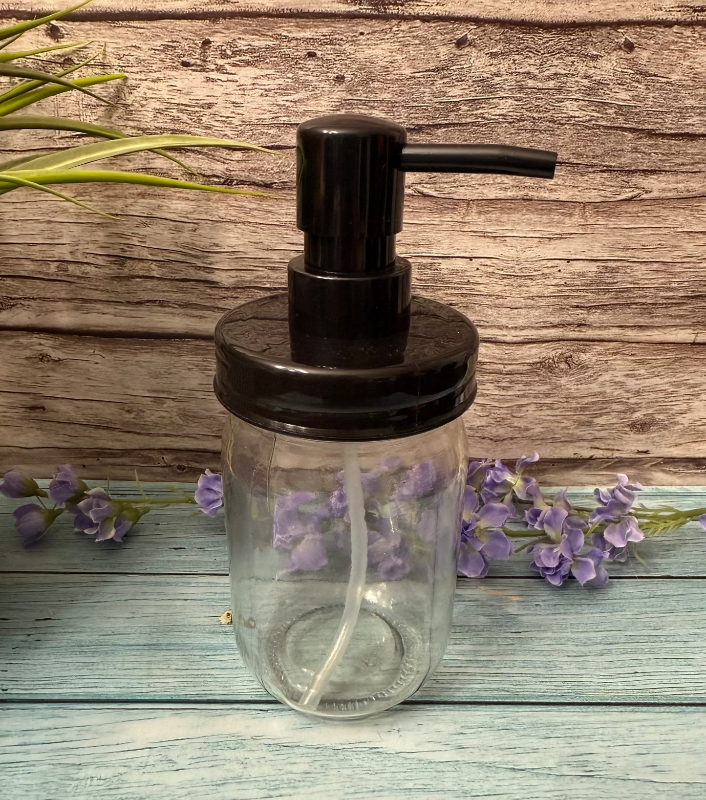 Clear glass soap dispenser with black pump on a wooden surface with purple flowers.